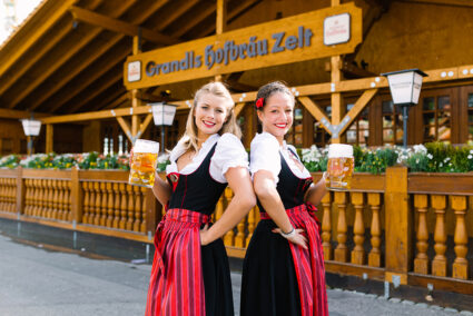 Zwei Frauen in Dirndl und mit einem Maß Bier in der Hand stehen vor dem Hofbräuzelt auf den Cannstatter Wasen